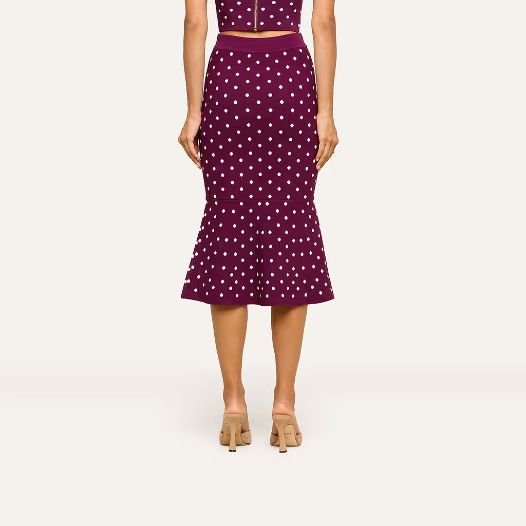 A model from the back wearing a high-waisted, flared midi skirt in deep purple with a white polka dot pattern, paired with beige heeled sandals against a light gray studio background.