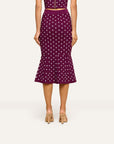 A model from the back wearing a high-waisted, flared midi skirt in deep purple with a white polka dot pattern, paired with beige heeled sandals against a light gray studio background.