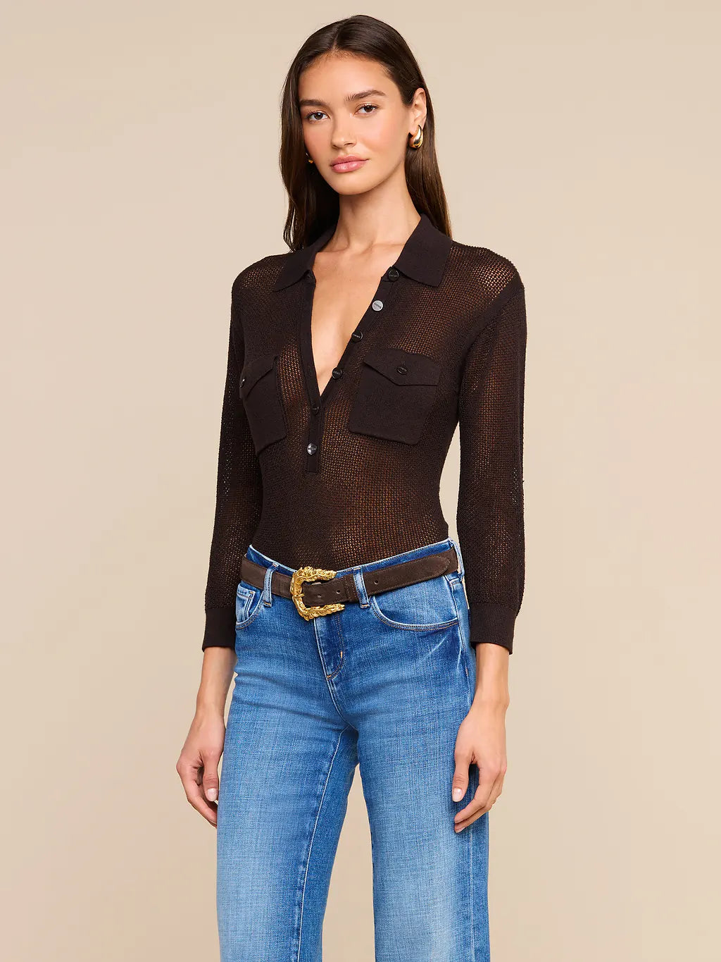 A woman modeling a dark brown sheer long-sleeve button-up shirt with chest pockets, paired with blue jeans and a brown belt, against a solid beige background.