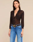 A woman modeling a dark brown sheer long-sleeve button-up shirt with chest pockets, paired with blue jeans and a brown belt, against a solid beige background.