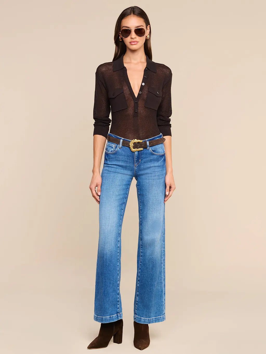 A model wearing a sheer black long-sleeve button-up shirt, blue flare jeans with a brown belt, and brown ankle boots, standing against a solid beige background.
