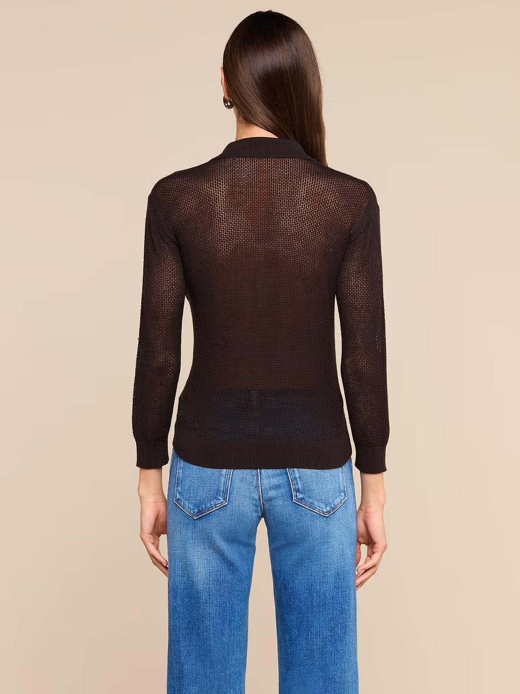 A woman viewed from the back wearing a dark brown sheer long-sleeve knit top and blue jeans, set against a solid beige studio background.