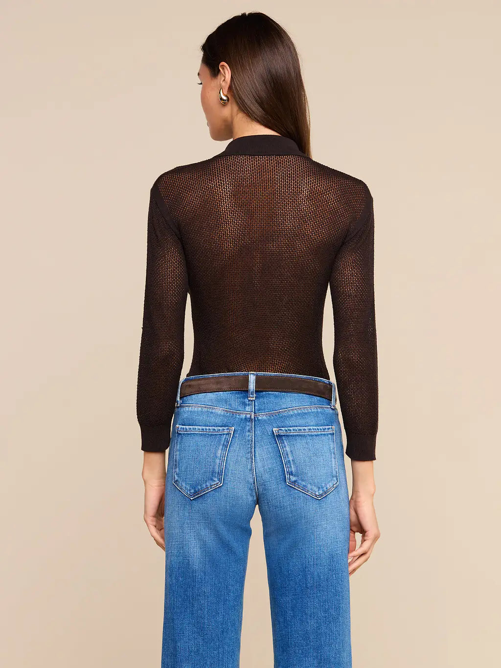 A woman shown from the back wearing a dark brown sheer long-sleeve mesh top and blue jeans against a solid beige studio background.