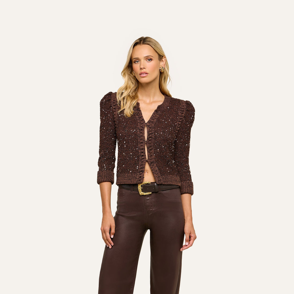 A woman modeling a dark brown cropped knit cardigan with textured sequins and puffed sleeves, paired with dark brown pants, against a solid off-white background.