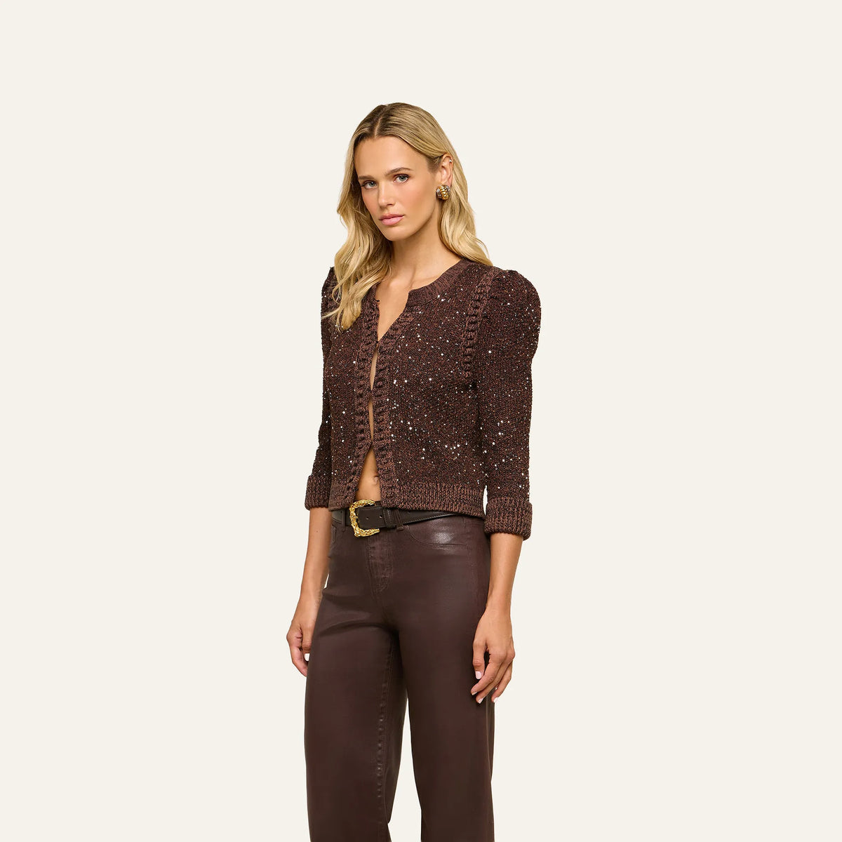 A blonde model wearing a dark brown textured knit cardigan with sequins and puffed sleeves, paired with dark brown pants and a gold belt, against a solid off-white background.