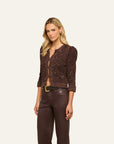 A blonde model wearing a dark brown textured knit cardigan with sequins and puffed sleeves, paired with dark brown pants and a gold belt, against a solid off-white background.