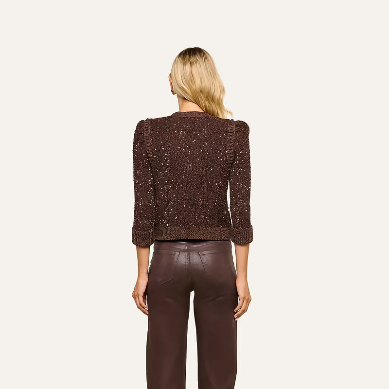 A woman viewed from the back wearing a dark brown textured knit cropped sweater with puffed shoulders and ribbed cuffs, paired with dark brown pants, against a plain off-white background.