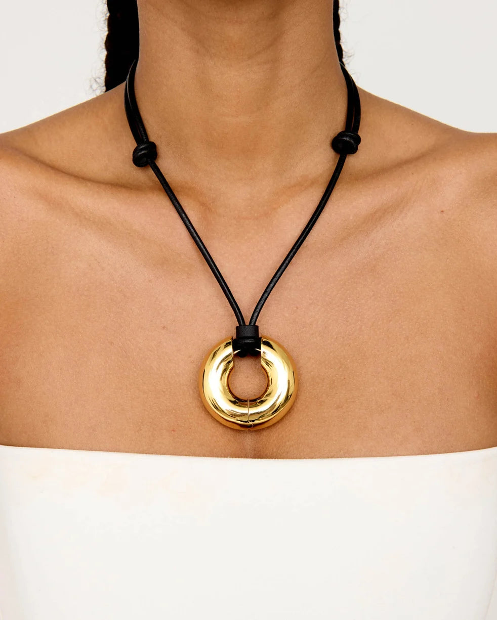 A close-up of a model wearing a black cord necklace with a polished gold circular pendant.