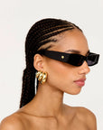 Woman wearing black sunglasses with a gold logo and gold hoop earrings, against a light gray background