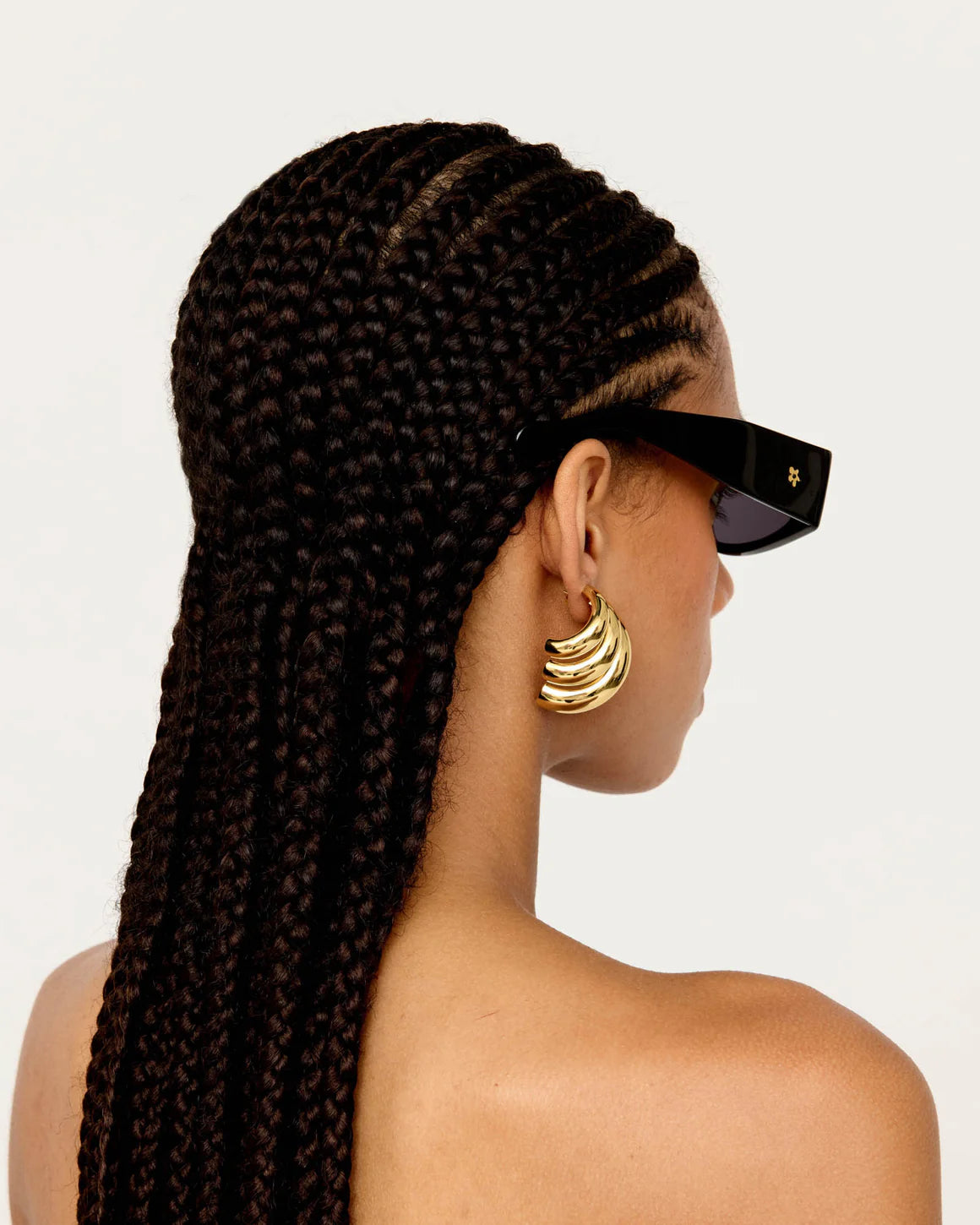 Woman with long braided hair wearing sunglasses and gold earrings on a plain background
