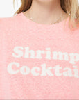 Person wearing a pink t-shirt with 'Shrimp Cocktail' text and red sunglasses on a white background