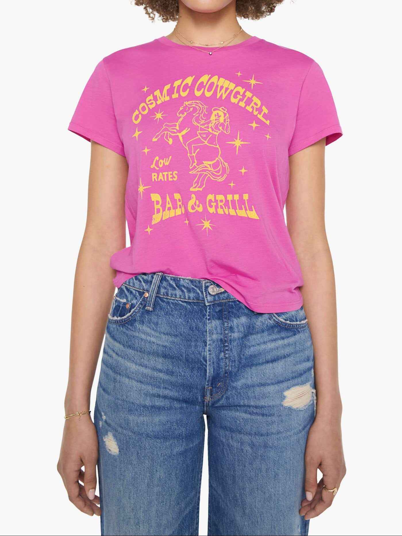A woman modeling a bright pink graphic t-shirt with a yellow retro cowboy illustration and text reading 'Cosmic Cowgirl Bar &amp; Grill'. She is wearing blue jeans and sunglasses against a plain white background.