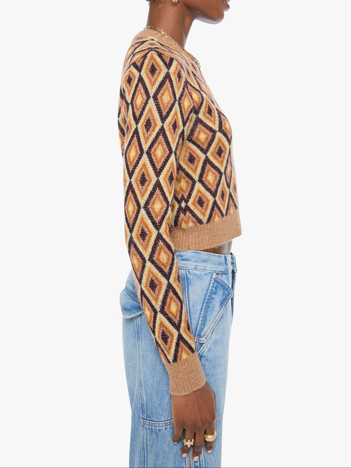 A side-profile view of a model wearing a cropped long-sleeve knit sweater with a geometric diamond pattern in tan, brown, and black, paired with light blue jeans against a plain white background.