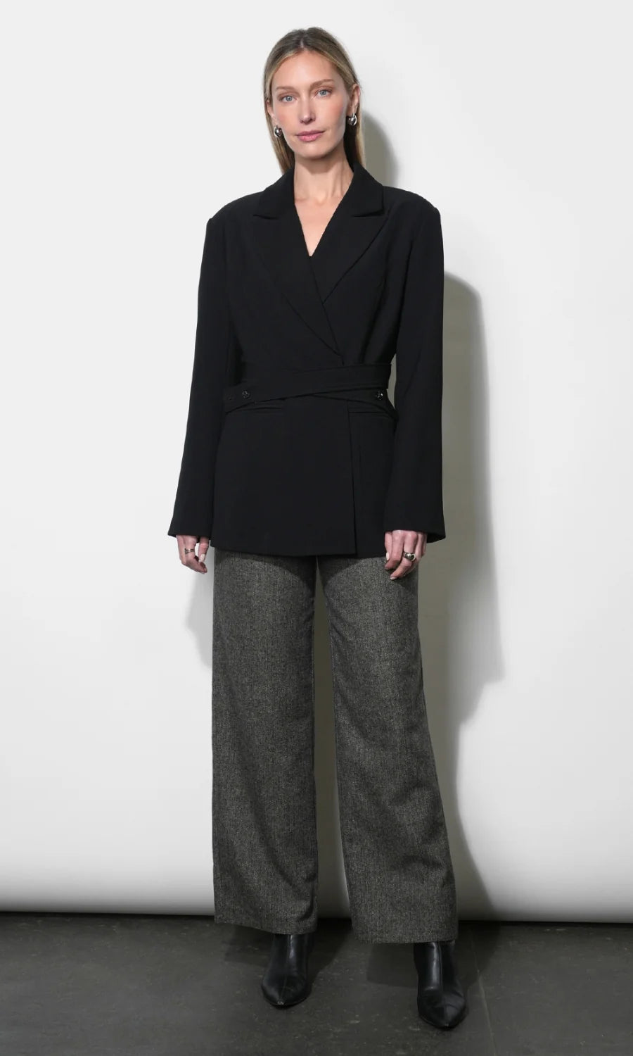 A model stands against a white studio wall wearing a black belted wrap blazer with wide long sleeves, paired with wide-leg charcoal gray trousers and black ankle boots.