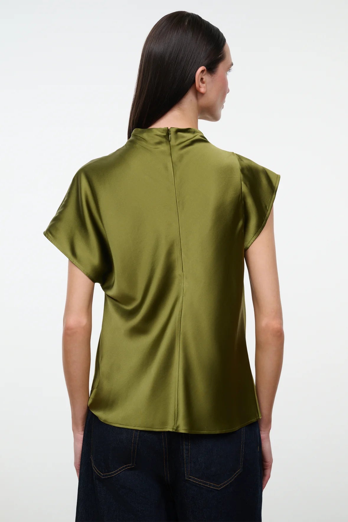 A woman viewed from the back wearing a short-sleeved olive green satin blouse and dark blue jeans against a plain white studio background.