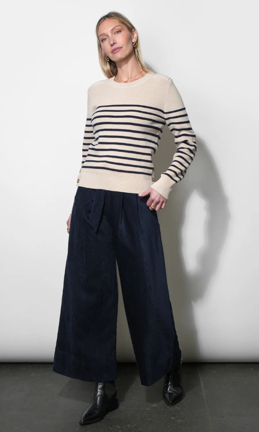 A woman modeling a cream and navy striped knit sweater with wide-leg navy trousers and black ankle boots, standing against a plain light gray wall.