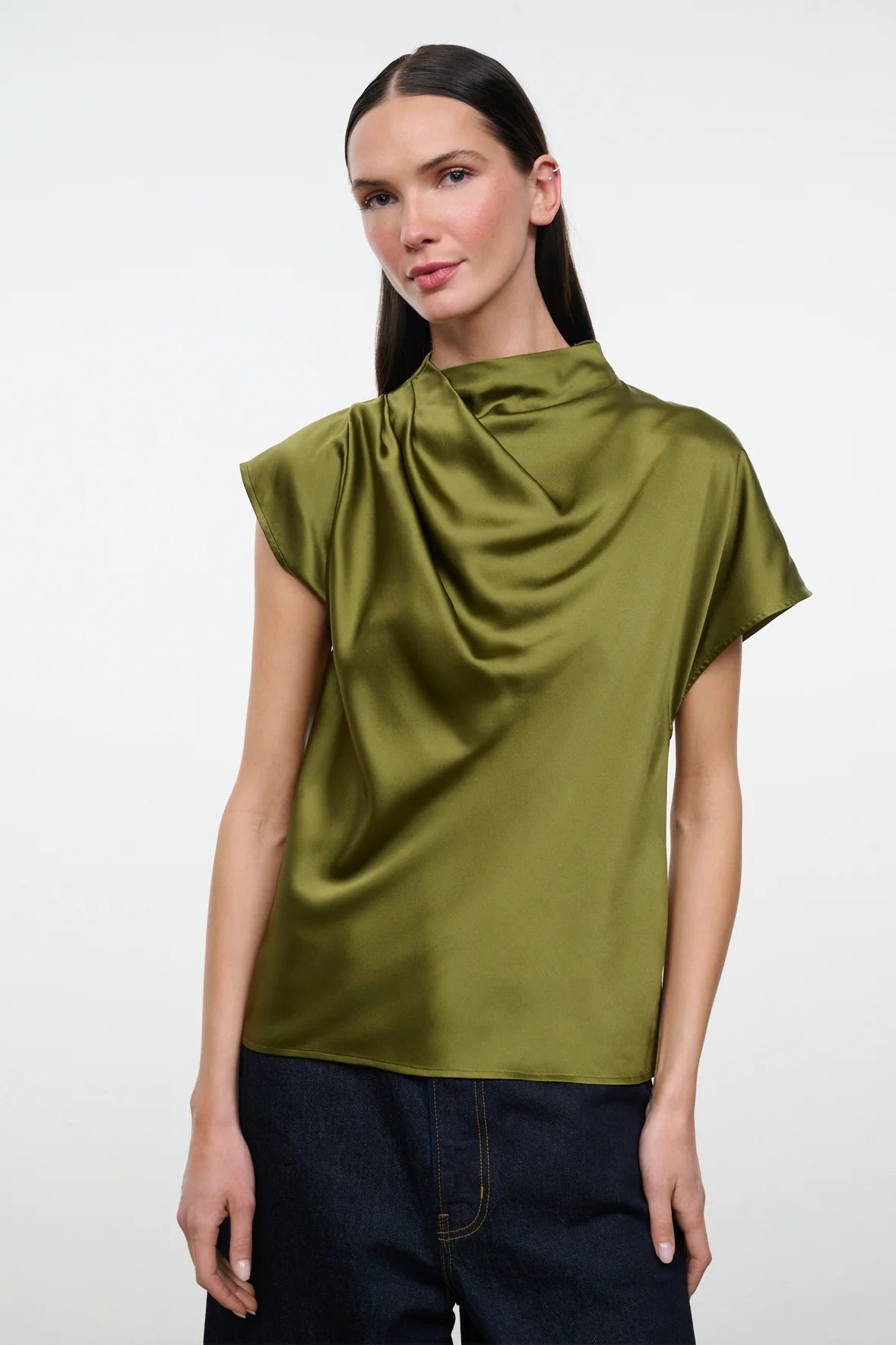 A woman modeling a short-sleeve olive green draped satin top with dark blue jeans, shot against a plain light gray studio background.