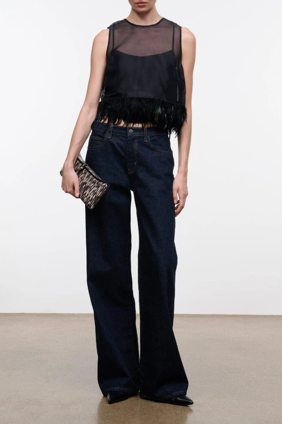 A model wearing a black sleeveless sheer mesh top with feather trim, paired with dark blue wide-leg jeans and holding a small patterned clutch, set against a plain white studio background.