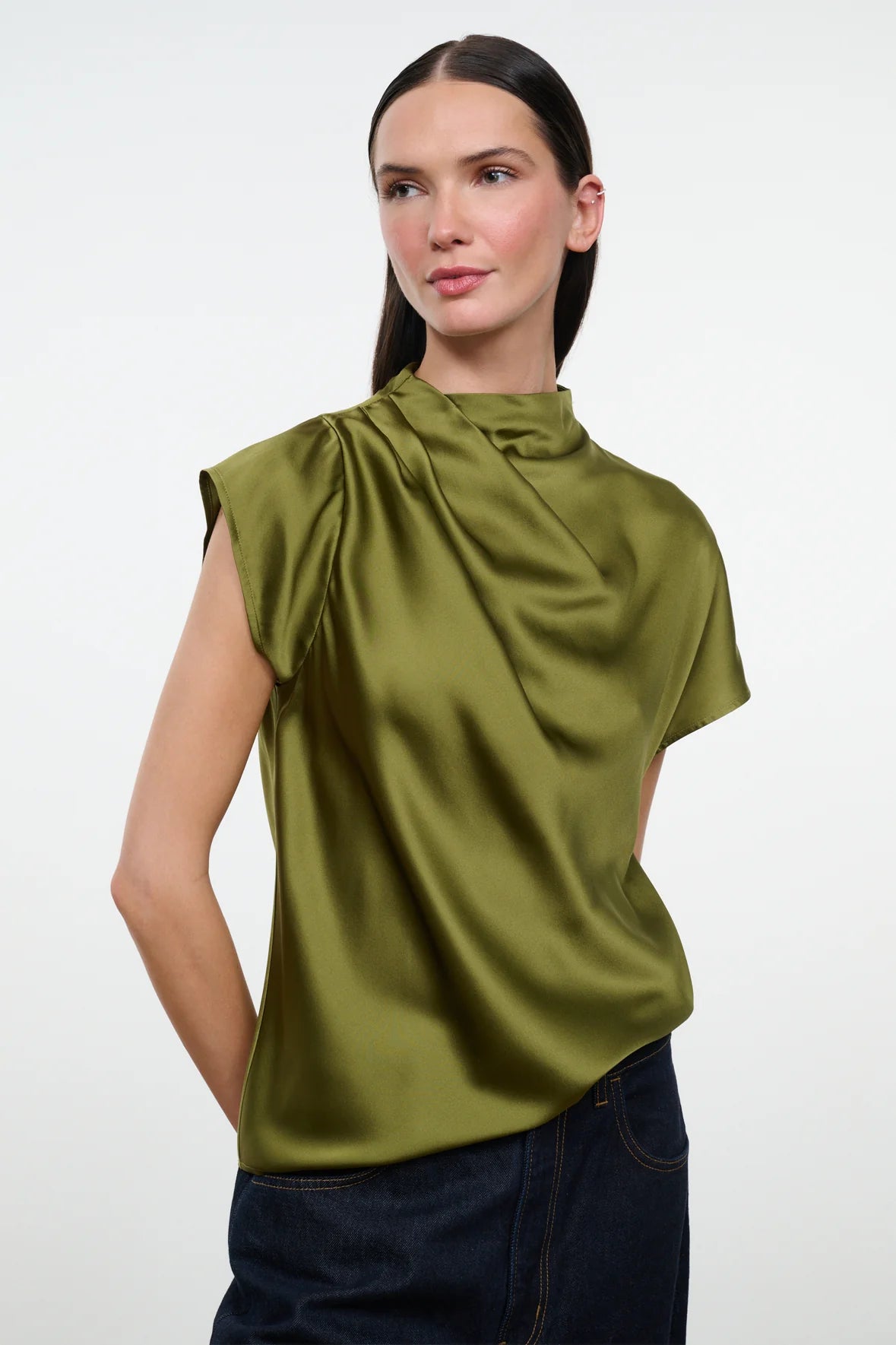 A woman modeling a sleeveless olive green satin top with a draped high neck, paired with dark blue jeans, against a plain light gray background.