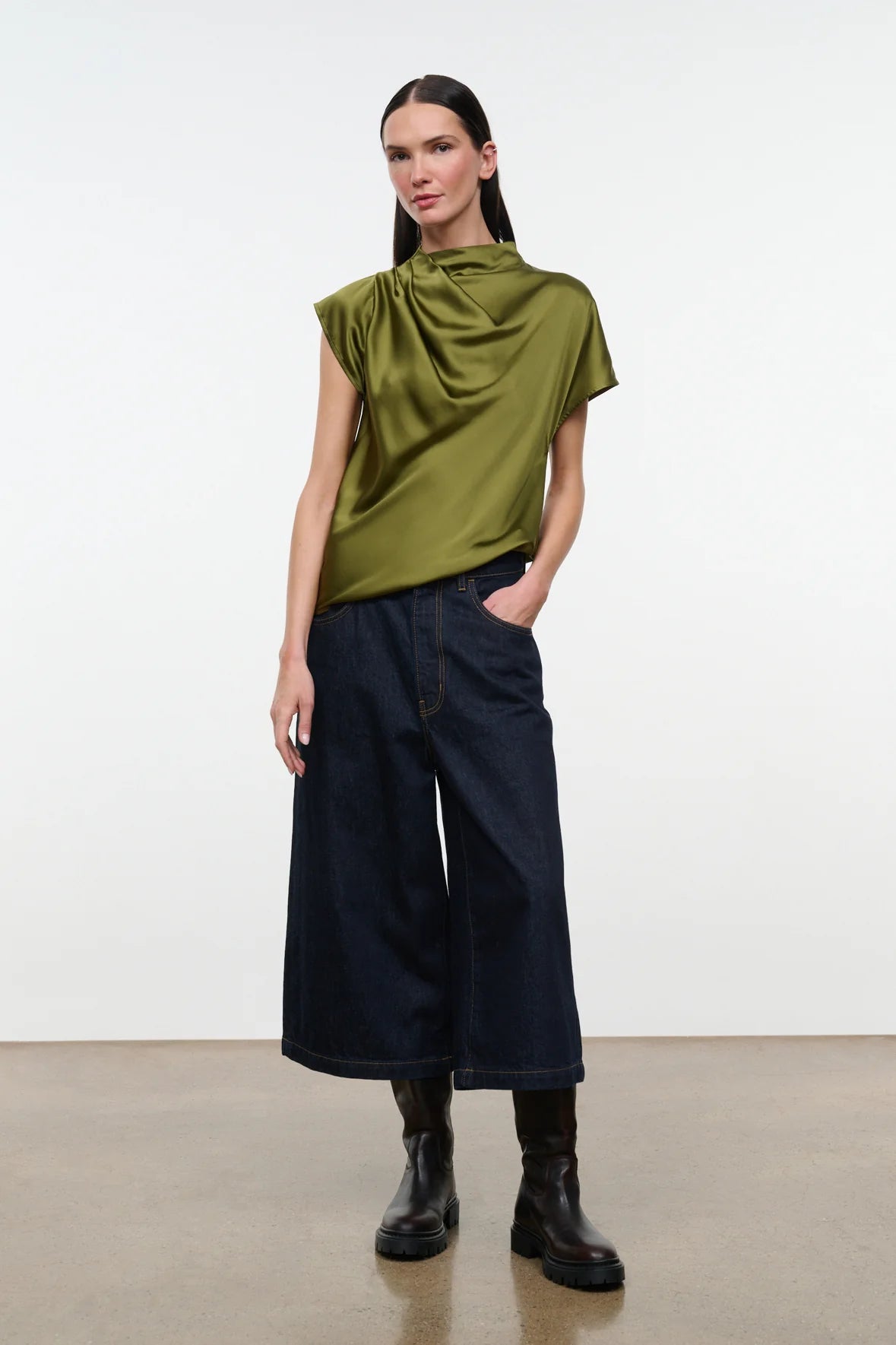 A model stands against a plain white studio background wearing a draped, short-sleeved olive green top, dark wide-leg denim pants, and black boots.