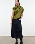 A model stands against a plain white studio background wearing a draped, short-sleeved olive green top, dark wide-leg denim pants, and black boots.