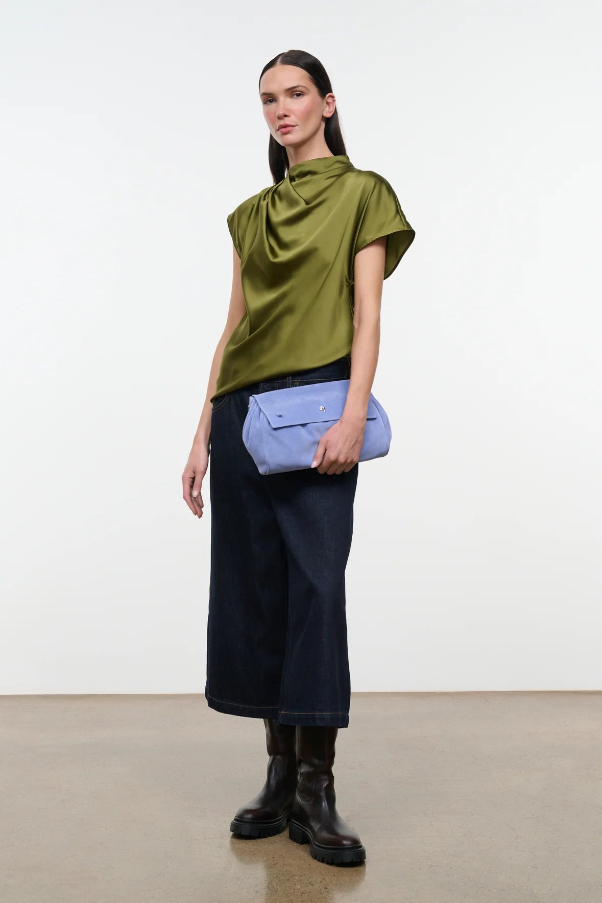 A model stands against a plain white studio wall wearing a draped olive green short-sleeve top, dark navy wide-leg cropped pants, black leather ankle boots, and holding a light blue envelope-style clutch.