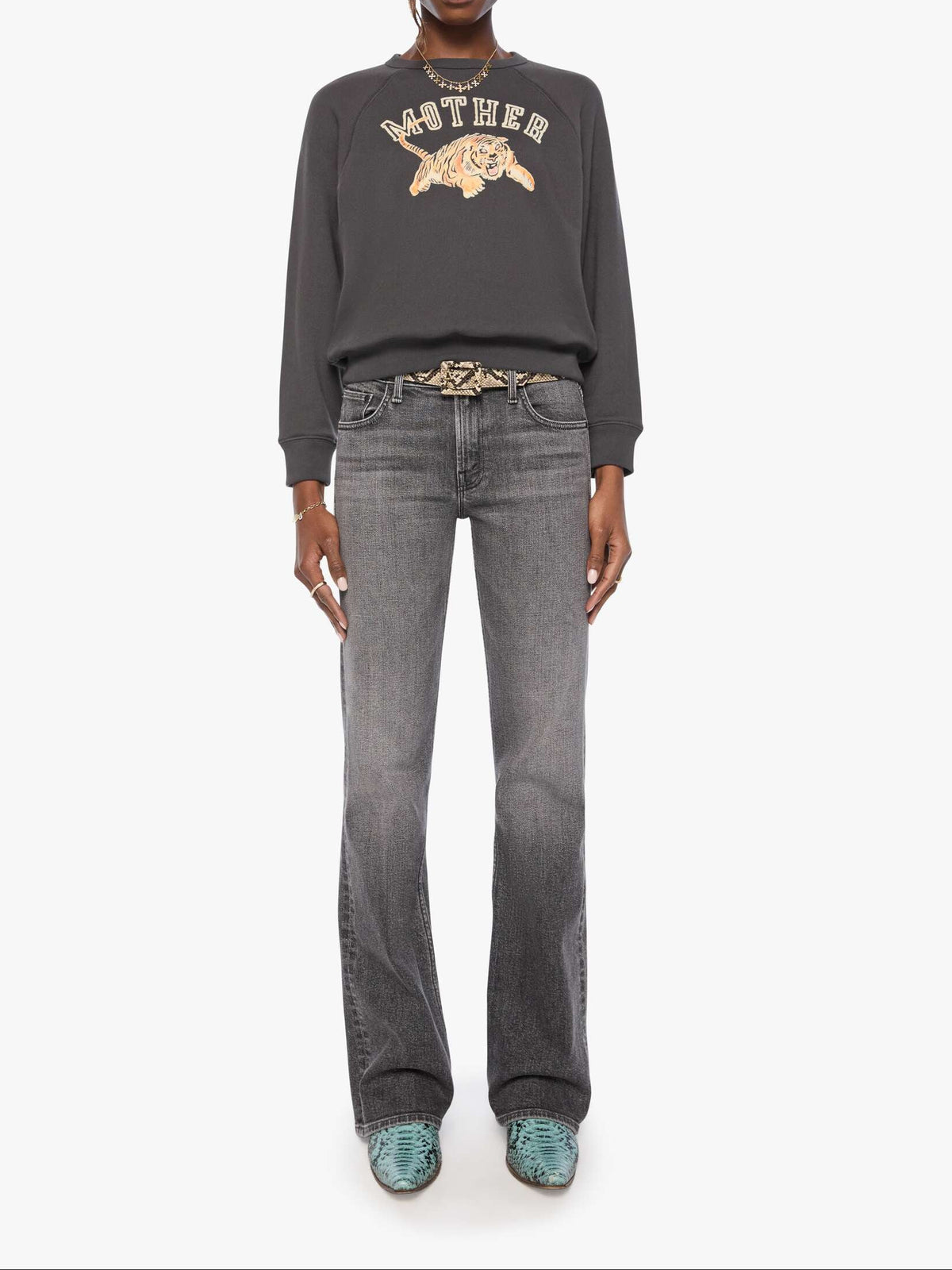 A model stands against a light gray studio background wearing a dark charcoal gray sweatshirt with a gold graphic and the word 'MOTHER' across the chest, paired with faded gray jeans, a decorative belt, and teal shoes.