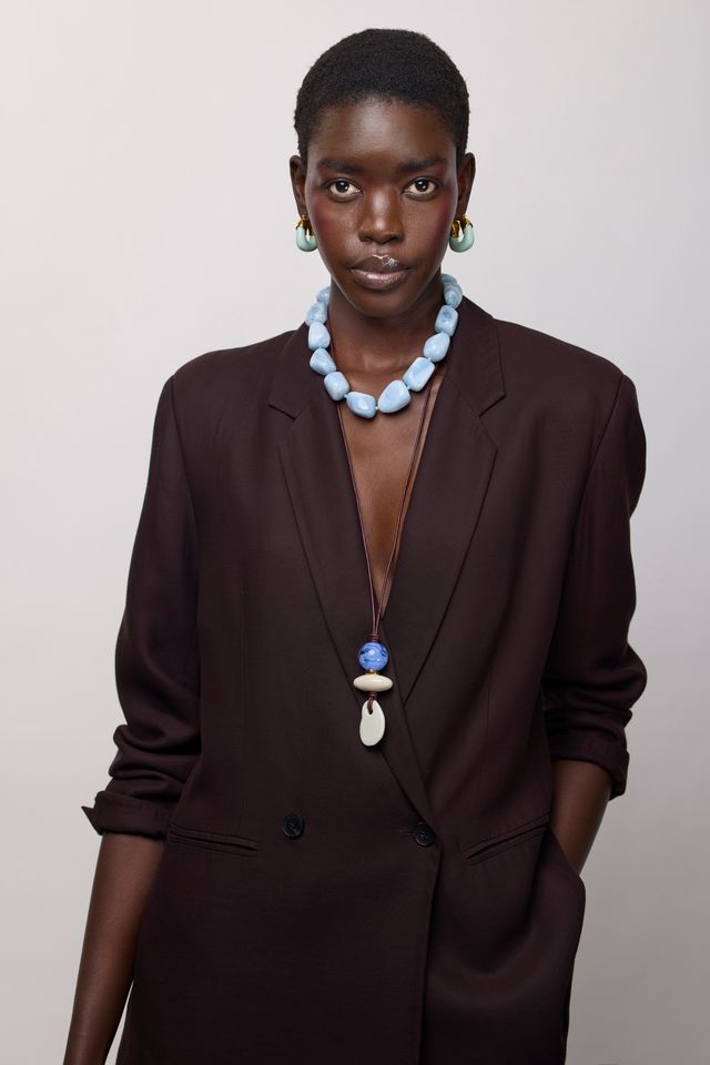 A model wearing a dark brown double-breasted blazer, a chunky light blue beaded necklace, and a long pendant necklace with blue and white beads, set against a plain light gray background.