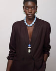 A model wearing a dark brown double-breasted blazer, a chunky light blue beaded necklace, and a long pendant necklace with blue and white beads, set against a plain light gray background.