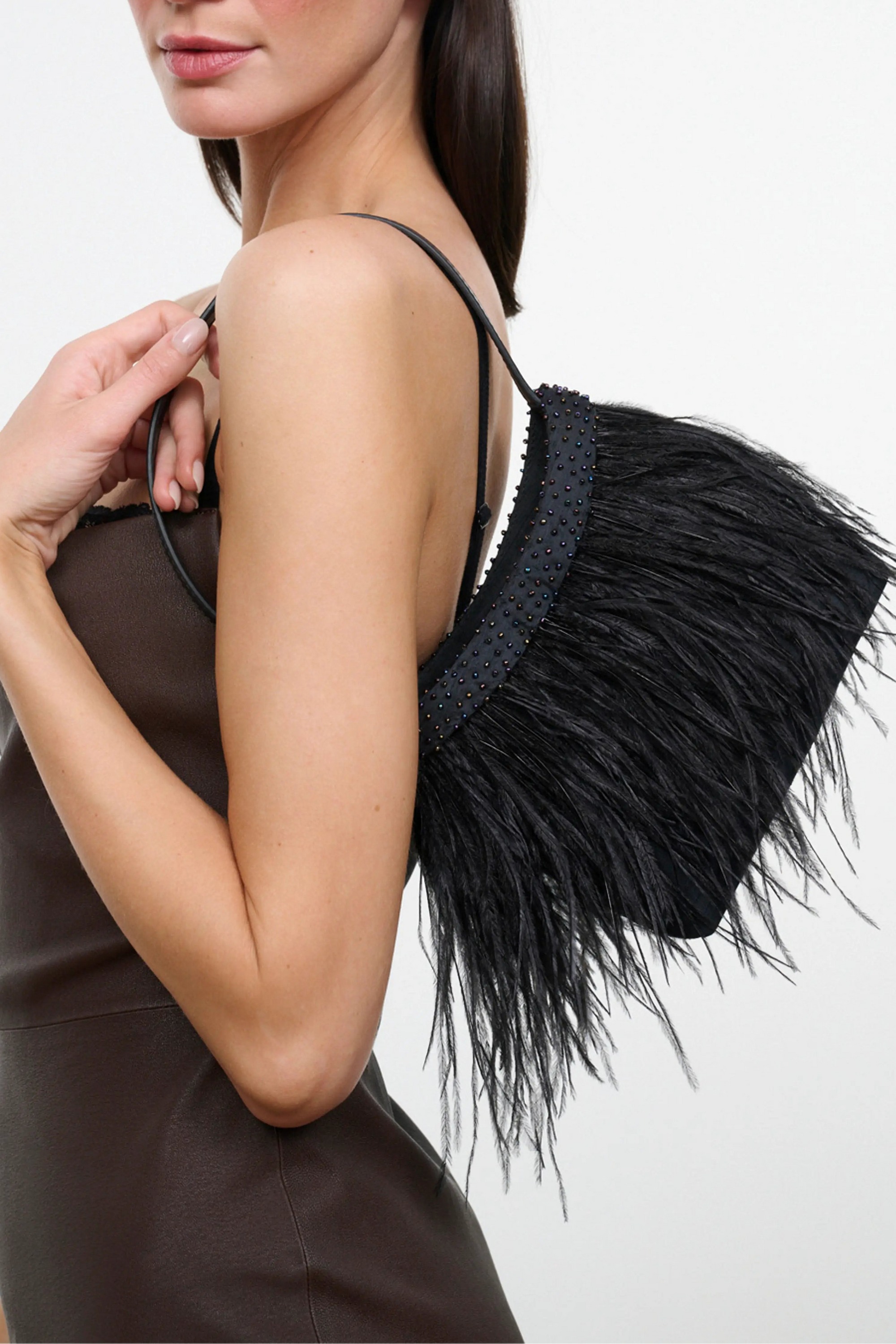 A woman modeling a black feathered shoulder bag with a dark brown strap, set against a plain light gray studio background.