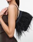 A woman modeling a black feathered shoulder bag with a dark brown strap, set against a plain light gray studio background.