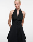 A blonde model wearing a black halter-neck mini dress with a button-front bodice and flared skirt, photographed against a plain light gray studio background.