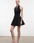 A model wearing a black halter-neck mini dress with a button front and flared skirt, paired with black and white patterned mule heels, standing against a plain white studio background.