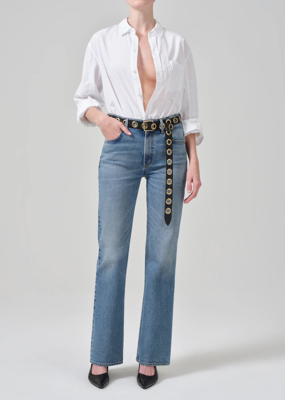 A model wearing a white button-down shirt, light-wash blue jeans with a decorative black belt featuring gold-tone circular hardware, and black pointed-toe heels, photographed against a light gray studio background.