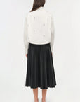 Person wearing a white jacket with black embellishments and a black pleated skirt on a white background
