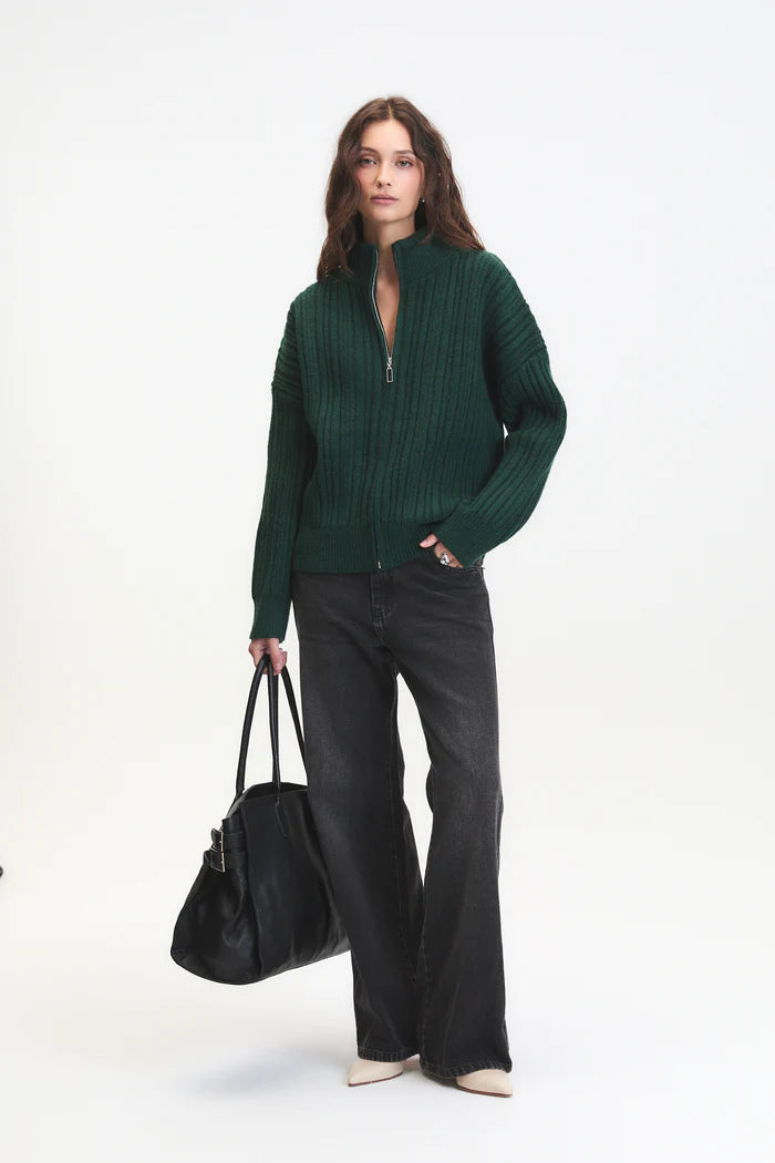 Woman wearing a green sweater and black pants, holding a black bag on a white background
