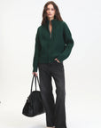 Woman wearing a green sweater and black pants, holding a black bag on a white background