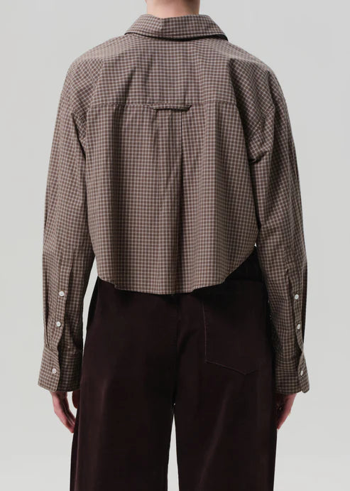 Person wearing a brown checkered shirt and dark pants on a light gray background, back