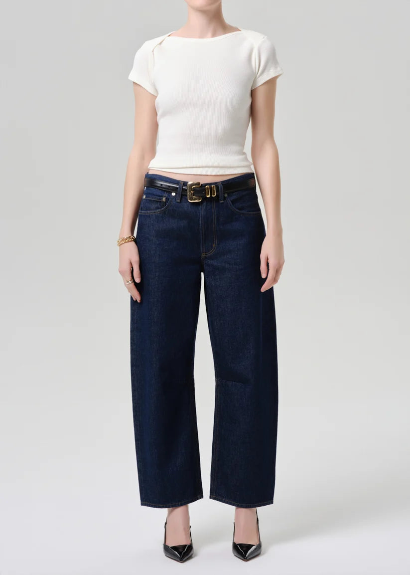 A model wearing dark blue wide-leg cropped jeans with a gold-tone belt, paired with a white short-sleeve top and black pointed-toe heels, against a light gray studio background.