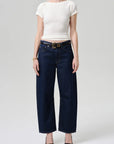 A model wearing dark blue wide-leg cropped jeans with a gold-tone belt, paired with a white short-sleeve top and black pointed-toe heels, against a light gray studio background.