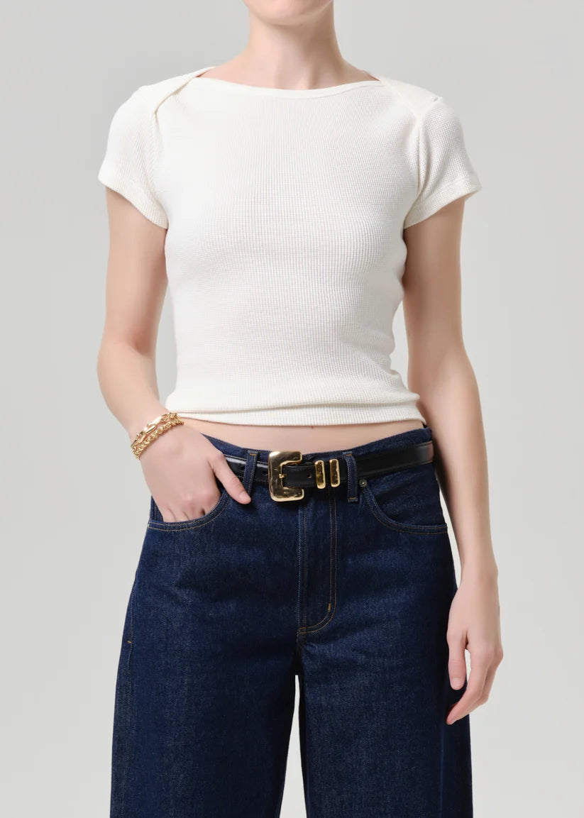 A model wearing a ribbed short-sleeve cream top and dark blue jeans with a gold belt, shown from the neck down against a light gray background.