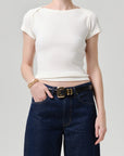 A model wearing a ribbed short-sleeve cream top and dark blue jeans with a gold belt, shown from the neck down against a light gray background.