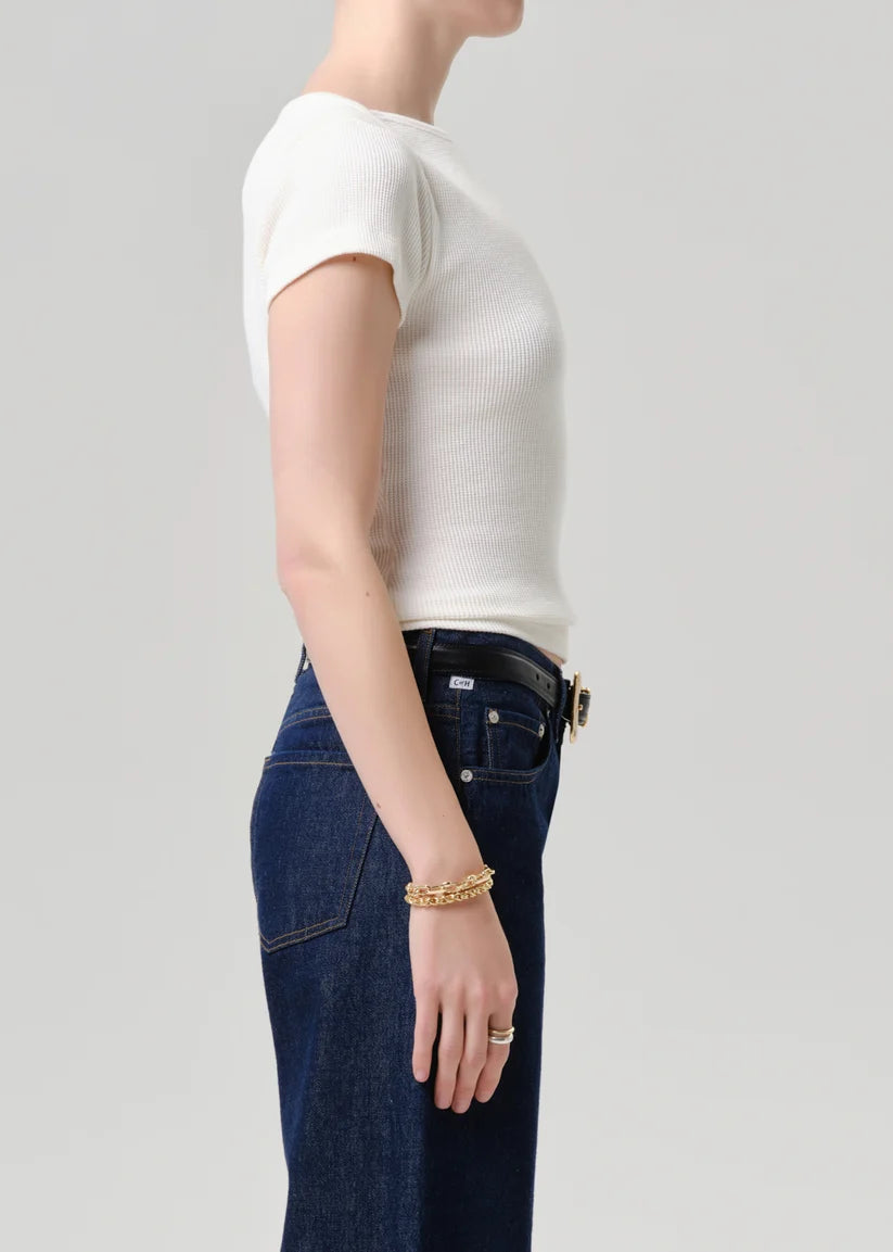 Side profile of a model wearing a white short-sleeve ribbed knit top tucked into dark blue jeans with a black belt, accessorized with a gold chain bracelet and ring, against a light gray studio background.