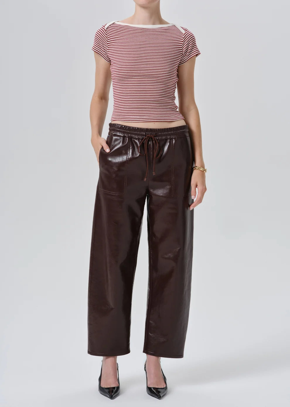 A model wearing a burgundy and white striped short-sleeve top with dark brown faux-leather wide-leg pants and black pointed-toe heels, photographed against a light gray studio background.