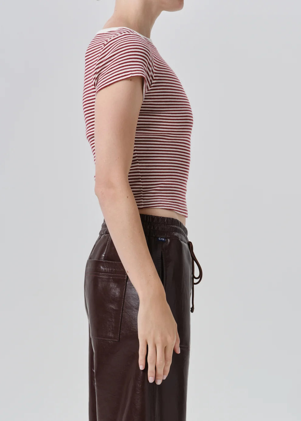 Side profile of a model wearing a short-sleeve cropped top with thin red and white horizontal stripes, paired with dark brown pants, against a light gray studio background.