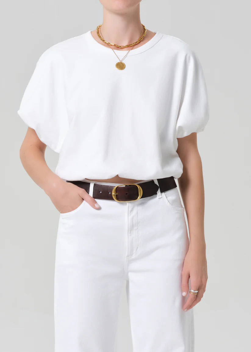 Person wearing a white top and white pants with a brown belt on a plain background