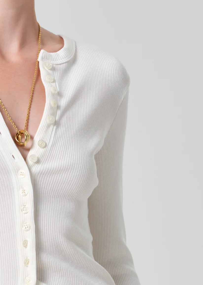 Close-up of a person wearing a white ribbed long-sleeve henley cardigan with cream buttons and a gold chain necklace with a double-ring pendant, against a light gray studio background.
