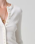 Close-up of a person wearing a white ribbed long-sleeve henley cardigan with cream buttons and a gold chain necklace with a double-ring pendant, against a light gray studio background.