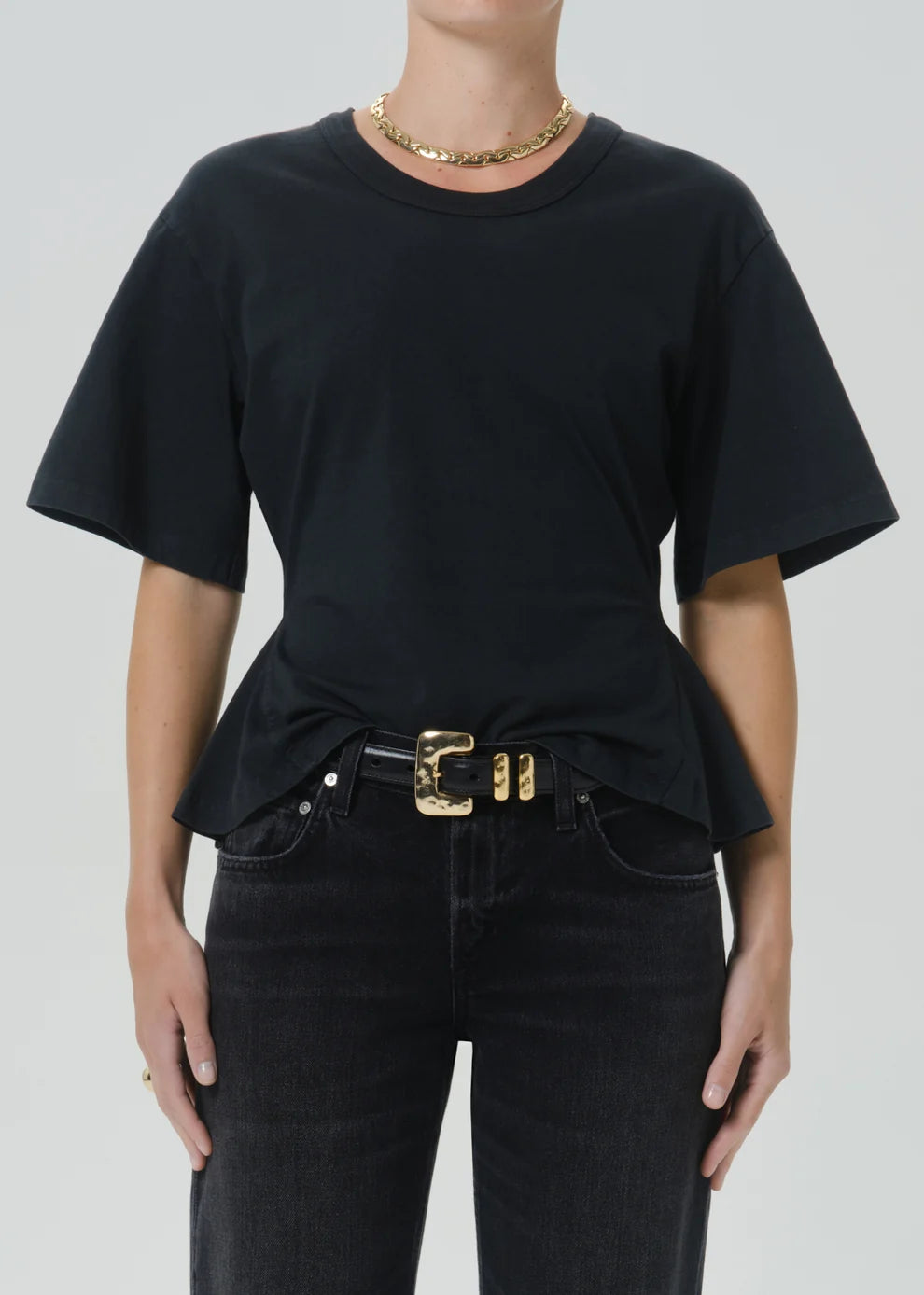 A model wearing a black short-sleeve crewneck t-shirt with a peplum hem, styled with a black belt featuring a prominent gold buckle, paired with dark jeans and a gold chain necklace.