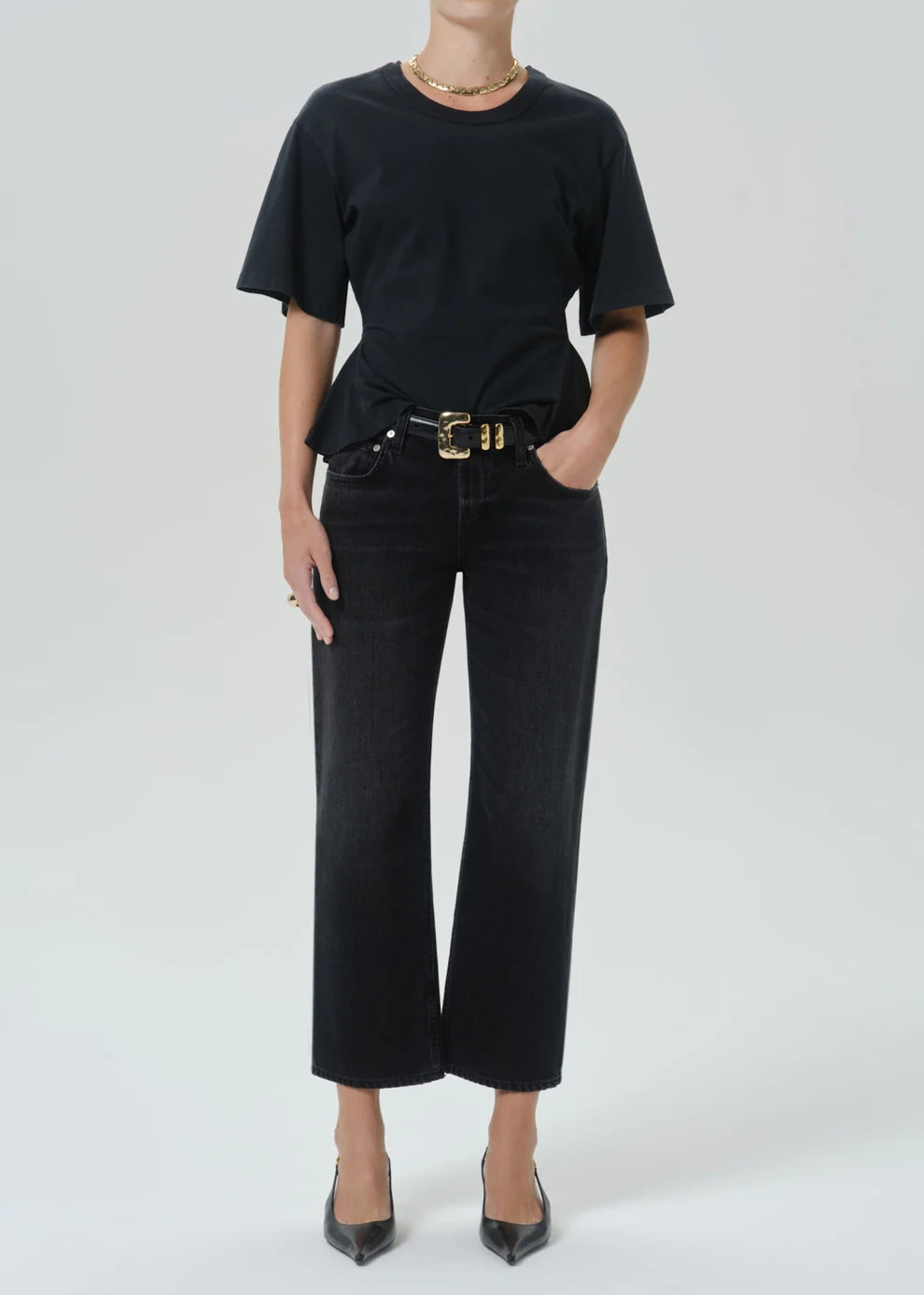 A model wearing a black short-sleeve crewneck t-shirt, black cropped wide-leg jeans with a gold-tone belt, and black pointed-toe heels against a light gray studio background.